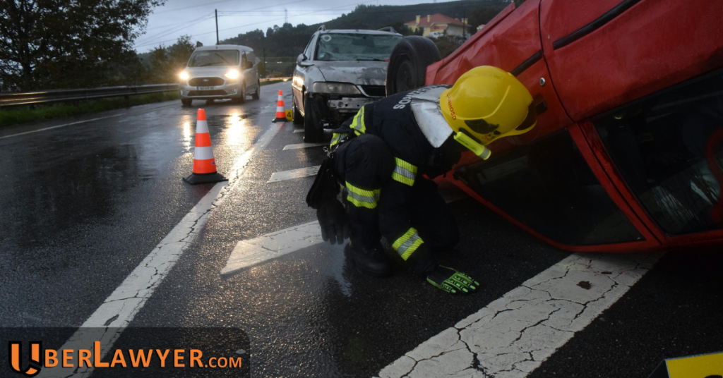 An emergency responder kneels beside an overturned car at a wet roadside crash scene, with traffic cones and a second damaged vehicle behind.